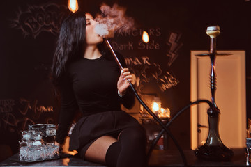 Sexy brunette girl in seductive black clothes smokes a hookah while sitting on counter in a nightclub or bar.