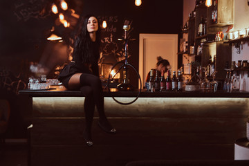 Naklejka na ściany i meble Seductive brunette girl in black clothes sitting with crossed legs on counter in a nightclub.