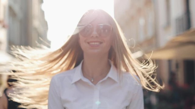 Charming young woman with a magnificent brown hair and stylish sunglasses. Attractive young lady is rushing in the city-center, she turns to camera and smiles