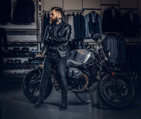 Brutal male dressed in black jacket posing with crossed arms near retro sports motorbike at the men's clothing store.