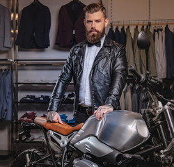 Stylish tattooed bearded man with dressed in black leather jacket and bow tie posing near retro sports motorbike at men's clothing store.