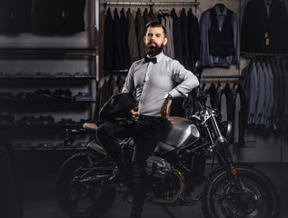 Fototapeta na wymiar Elegantly dressed tattooed male with stylish beard holds black leather jacket posing near retro sports motorbike at the men's clothing store.