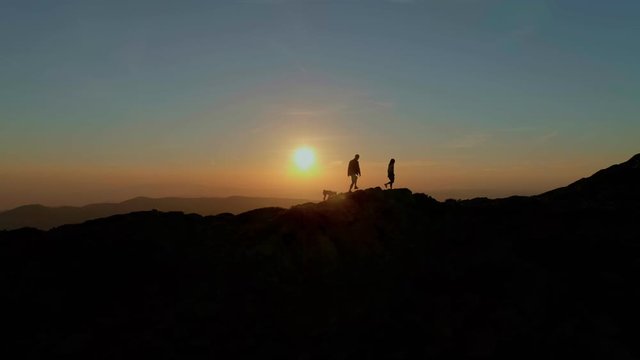 Aerial drone footage of couple and trail dog walk on edge of crest of mountain during beautiful sunset, figure silhouettes adventures and exploration of outdoors
