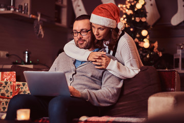 Portrait of an adorable couple - charming woman in Santa hat hugging her man and using a laptop.