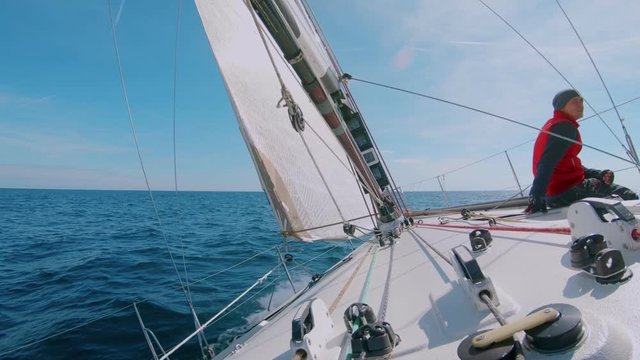 Slow motion wide beautiful shot of man or professional sailor or amateur sea lover sit on deck of sailboat under big white sail, during race in open ocean on warm sunny summer day