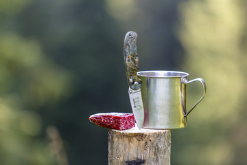 Close-up composition of folding pocket knife stuck vertically in tree stump, sausage and tin mug...