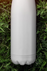 Stainless thermos water bottle, white color. Mockup isolated on green grass background with sunlight effect. Glossy aluminum vacuum thermo white tumbler flask.