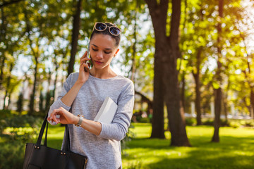 College student talking on mobile phone in park. Young woman holding book and checking time
