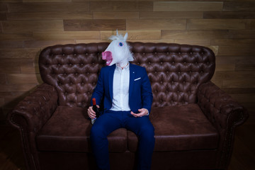 Funny unicorn in elegant suit sits on sofa with bottle of wine. Unusual man with headache at home....