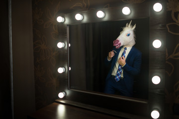 Glamour guy in elegant suit and comical mask looks at himself in the mirror in dressing room. Freaky young man in stylish room posing like a boss. Unusual unicorn.