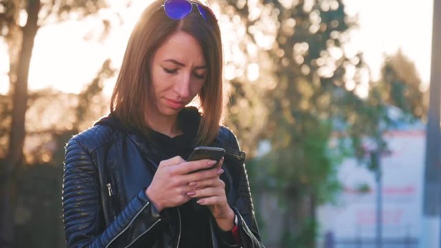 Attractive young girl stands in middle of busy buzzling city on summer sunset. Gorgeous young woman in a bright sunlight uses her phone.