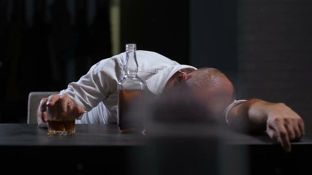 Portrait of alcoholic adult businessman holding glass of whiskey sleeping drunk on the table in domestic interior. Wasted drunk man in midlife crisis lying on the table in alcohol abuse and alcoholism