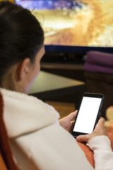 young woman looks at her smarthphone with white screen, sitting on the sofa, in front of the tv