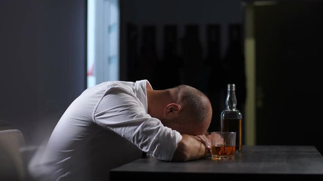 Depressed man in alcohol abuse experiencing midlife crisis and business failure lying on the table at home. Wasted drunk businessman sleeping at desk with bottle and glass of whiskey in front of him.