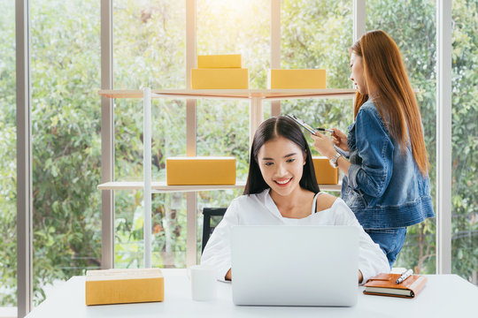 Business owner concept. Young Asian woman smiling happy using laptop working and checking customers order products packaging in modern home office with brown package box in hands