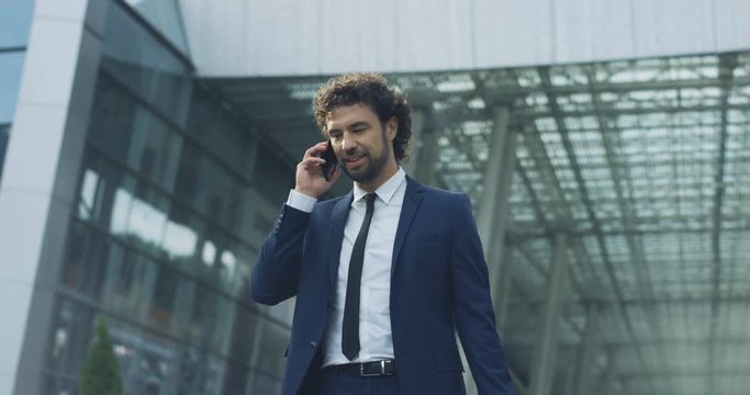 Caucasian young attractive man in the suit and tie standing outside big urban building and answering his moile phone ringing with a smile.