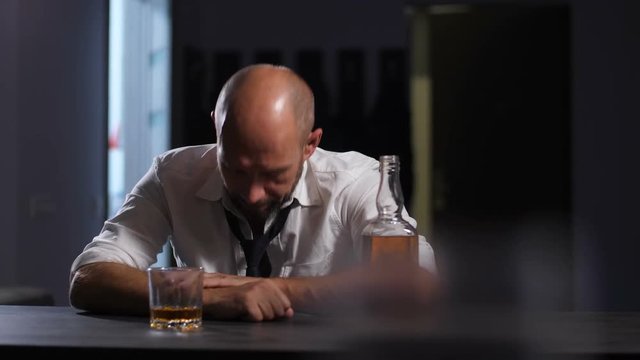 Sad mature man in shirt and tie feeling depressed while drinking alcohol at home after work. Male executive putting head on hands. Whiskey bottle and glass on the table. Dolly.