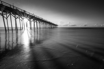Black & white view of sun rising on the horizon behind a pier