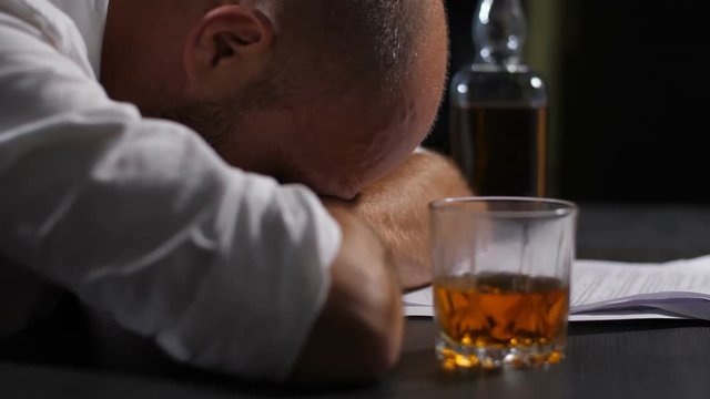 Close-up of overworked hopeless businessman leaning his head on hands with notifacation of company bankruptcy lying on the table. Depressed man avoiding problems in alcohol abuse indoors. Dolly shot.