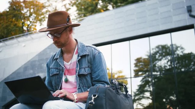 Portrait of young bearded hipster man and in hat sits on bench in city center in park using laptop, slow motion