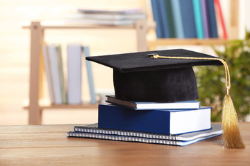 Graduation hat with books and notebooks on table against blurred background