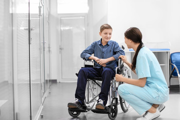 Young female doctor taking care of little boy in wheelchair indoors