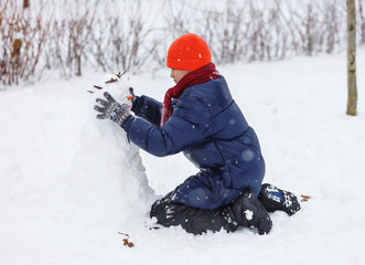 Pretty boy in warm blue jacket, red hat makes a snowman at winter day. Winter activities, fun