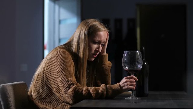 Jilted attractive wife with glass of red wine sitting in loneliness and crying at home after divorce. Stressed sad adult woman in deep depression and alcohol abuse feeling sorry for herself indoors