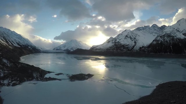 Wide aerial, snow capped mountains over frozen river at sunset