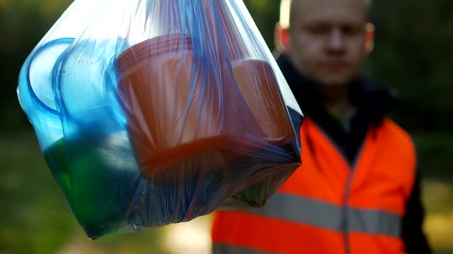 A man in a signal orange vest holds a package with garbage on the background of nature, forest, close-up, garbage collection, a janitor, volunteer, rubbish