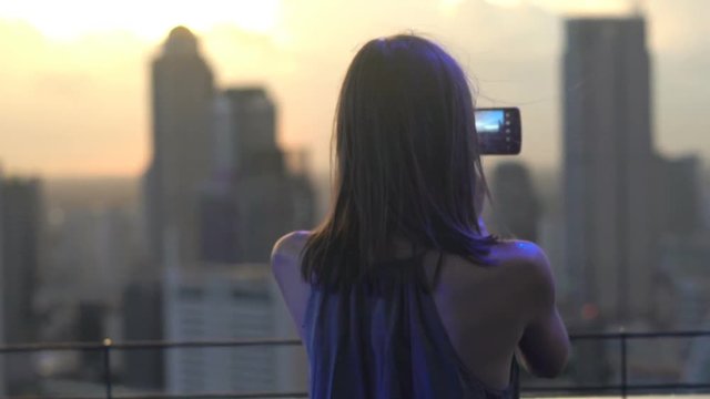 Young woman taking photo of cityscape with cellphone in skybar during sunset
