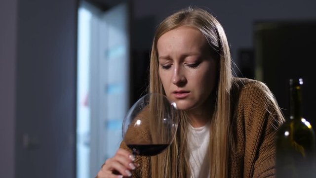 Full-fledged alcoholic woman holding glass of wine in trembling hands and sipping alcohol while sitting alone in dimmed room. Hopeless destroyed female wasting her life and beauty in alcohol abuse.
