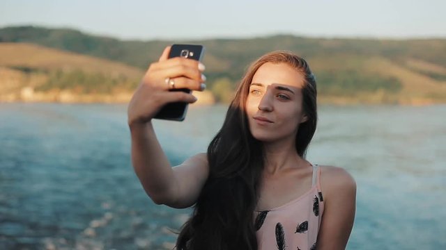 A beautiful girl making selfie with her phone while standing in front of the river and mountains. Half-body portrait