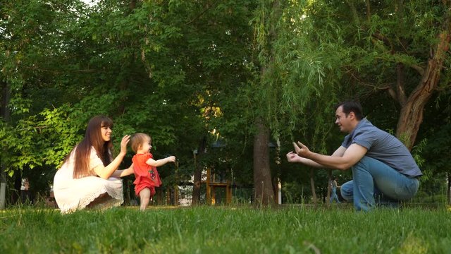 mom and dad learn to walk the baby, child takes first steps on lawn in park in summer