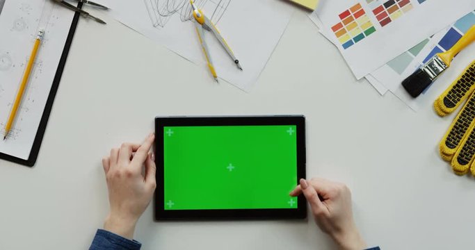 Close up of the top view on the female hands scrolling and tapping on the chroma key screen of the black horizontal tablet device on the designer desk with drawings stuff. Green screen. Tracking