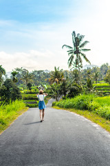 Fototapeta na wymiar Tropical portrait of young happy woman with straw hat on a road with coconut palms and tropical trees. Bali island.