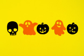 Holiday decorations for Halloween. Orange paper ghosts and black paper pumpkins and skull on a yellow background, top view.