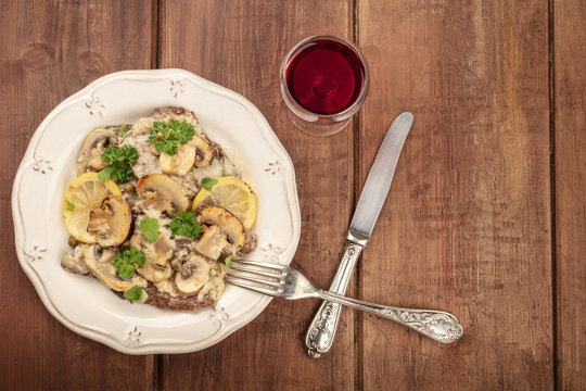 Overhead photo of scallopine di vitello, veal scallopini, with a mushroom sauce, lemons, and parsley, on a dark rustic texture with red wine and copy space