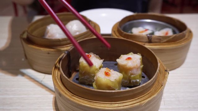 4K Using chopsticks eating Siu mai or Shao Mai (steam minced pork shrimp dimsum in yellow wonton wrapper) with background of other dimsum like Har Gow, pork wrap with Seaweed  left in bamboo baskets.
