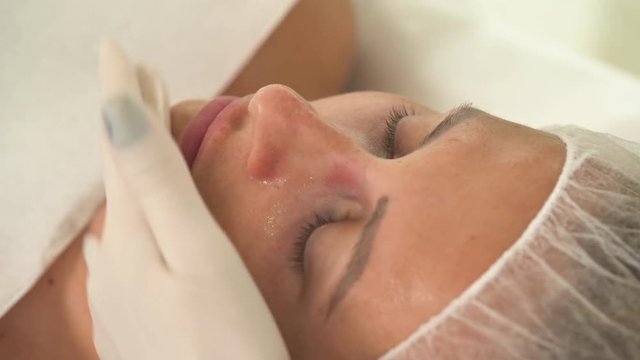 A top view of a woman's face after cleansing and mask application. A hand in a white rubber glove applies some gel on the face in strokes.