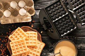 Wooden board with tasty waffles, maker machine and bowl with batter on dark wooden table