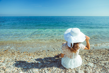 The back view of young beautiful woman in white dress sitting on the beach and looking at the sea...