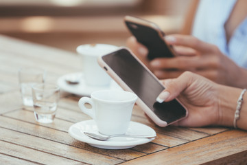 Close-up of female hands holding phones chatting on social network or shopping online in cafe with two cups of italian espresso coffee. Compulsive smartphone and internet use (addiction) in restaurant