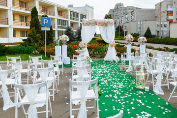 Beautiful decoration of the wedding ceremony. White wooden chairs for guests on either side of the walkway, which leads to festive arch decorated with flowers
