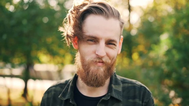 Slow motion portrait of happy bearded guy hipster in military style clothing standing in the park smiling and looking at camera. People and emotions concept.