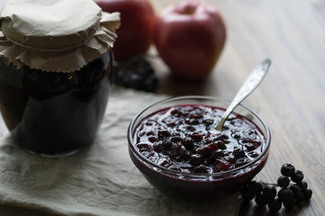Home made black chokeberry and apple confiture in a glass jar on a wooden background.