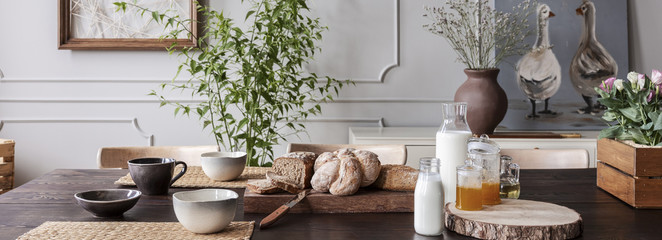 Panorama of wooden table with bowls, bread, milk and honey in grey dining room interior. Real photo