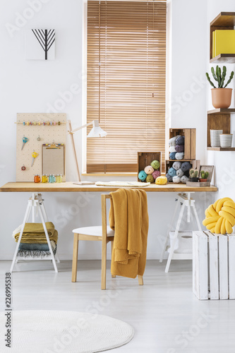 Blanket on chair at desk with yarns in white home office interior with blinds at windows. Real photo