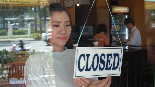 Young Asian businesswoman turning sign from close to open on glass entrance door of her bar and welcoming guests, traffic seen in reflection
