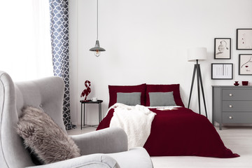 A glamour bedroom interior in white, gray and burgundy with a bed, an armchair, a drawer cabinet and lamps . Real photo.